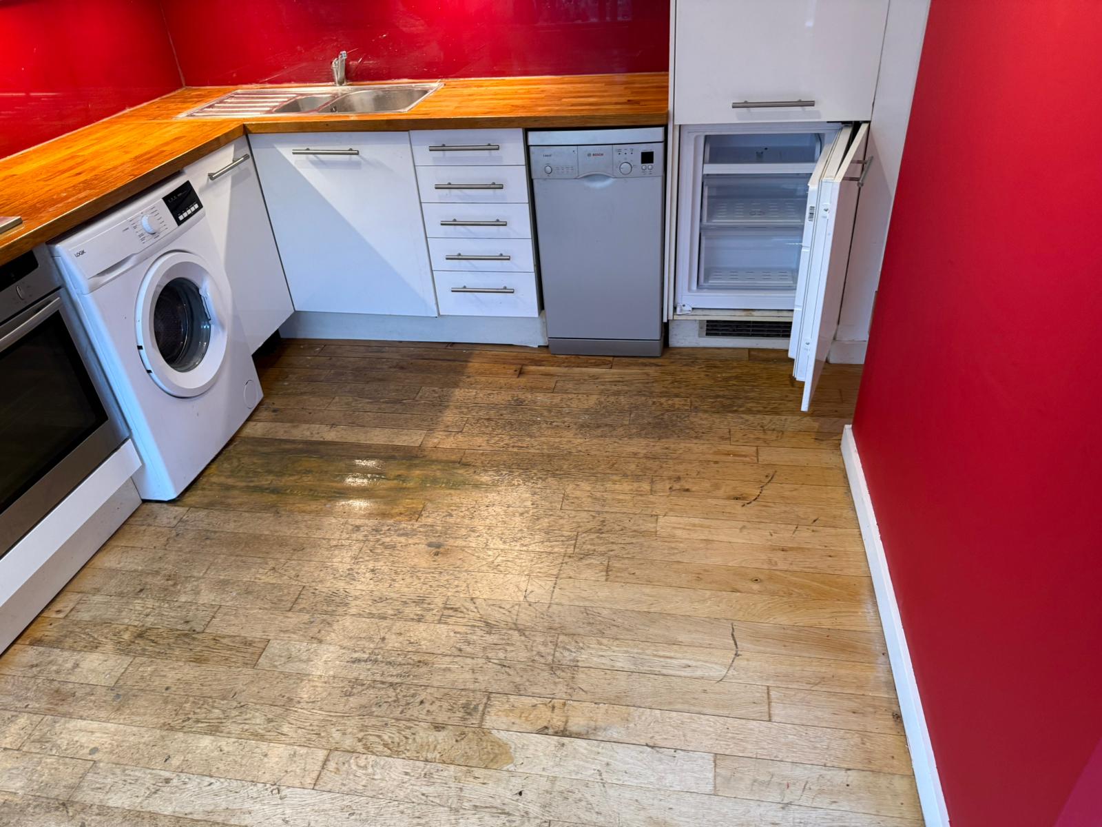 A worn and heavily scratched wooden kitchen floor, showing stains and discoloration around the washing machine area.