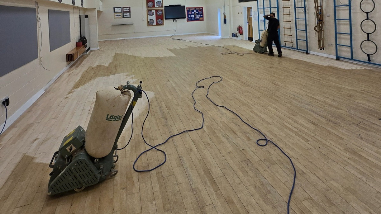 Hardwood Floor Refinishing in Southwark – maple strip hardwood school hall floor being sanded with large commercial machines, showing mid-refinishing progress across the gym area.
